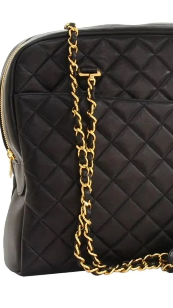 A close-up product photograph from 2_5.webp showcasing the classic styling of a black quilted leather bag. The bag features a detailed diamond-pattern quilting and a gold zipper closure visible on the left edge. A prominent shoulder strap made of a polished gold metal chain intertwined with a strip of black leather drapes vertically across the center of the bag, highlighting the combination of textures.