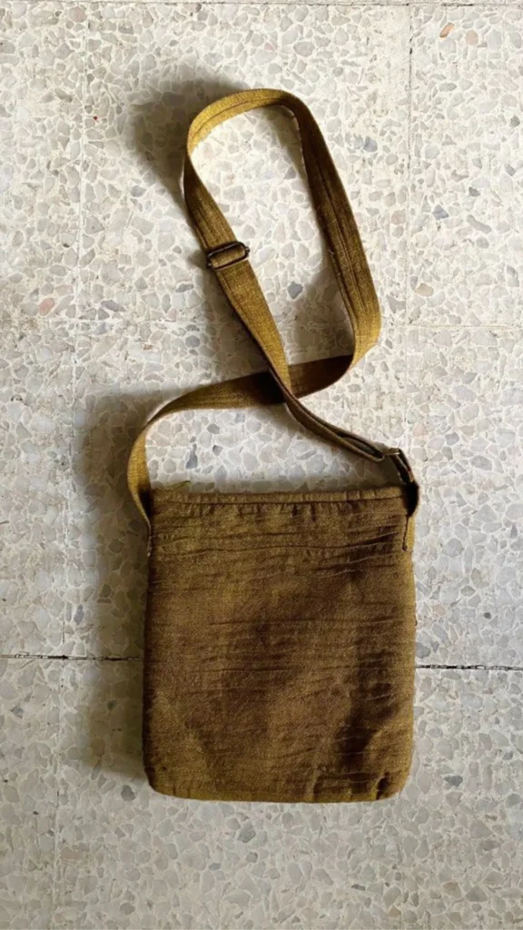 A flat-lay view from 2_6.webp featuring a minimalist, rectangular crossbody bag resting on a speckled terrazzo tile floor. The casual bag is made of a textured fabric in an earthy olive or moss green shade, showing subtle horizontal crinkle lines across the body. It features a matching wide fabric shoulder strap with a metal adjustment slider looped above the bag, emphasizing a simple, functional aesthetic.