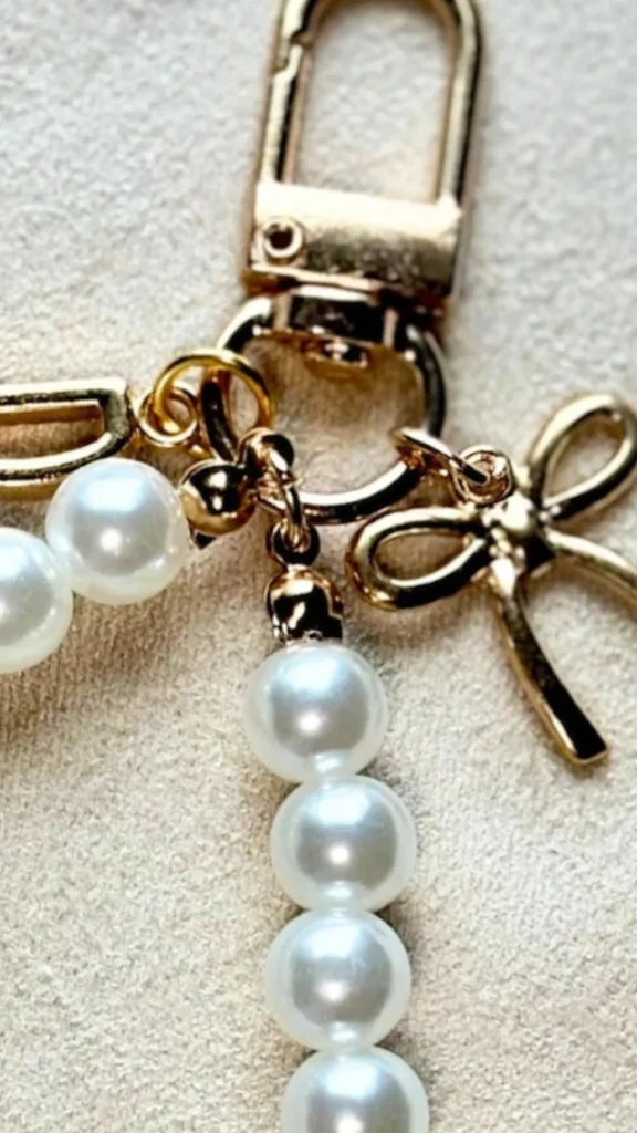 A close-up product photograph from 2_8.webp showing a bag charm or strap accessory made of white faux pearls and gold details. A large, polished gold-toned lobster claw clasp serves as the main attachment point. Connected to it by small gold jump rings are strands of glossy white pearls, alongside a dainty, decorative gold bow charm. The elegant accessory rests on a soft, textured beige fabric surface.