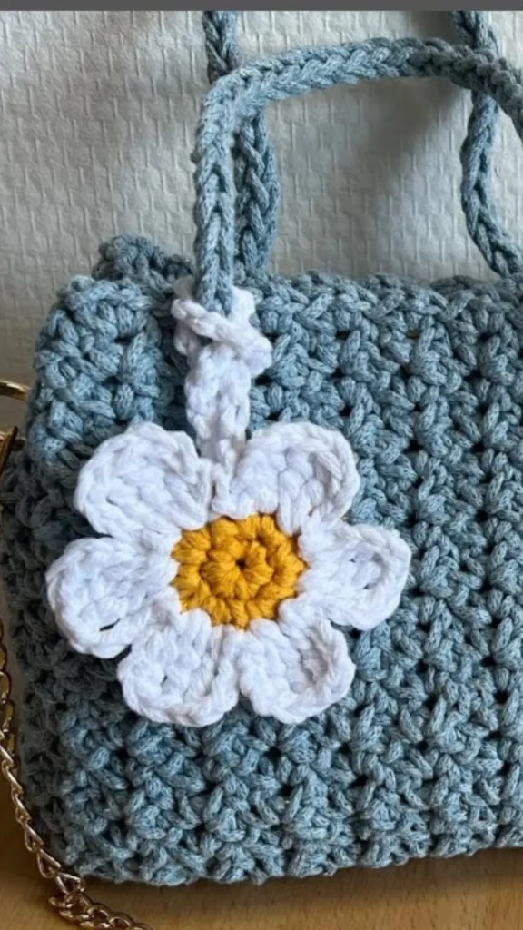 A close-up product photograph from 2_9.webp showcasing a handmade crochet daisy flower charm attached to a coordinating bag. The charm features a bright yellow crocheted center surrounded by six white yarn petals, loop-tied to the handle base with a white crochet yarn strap. It hangs against a chunky, textured dusty-blue crocheted tote bag that includes braided top handles and a gold chain shoulder strap.
