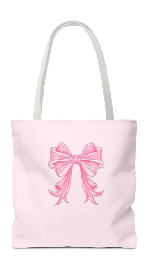 An image from file "2_20.webp" featuring a studio shot of a pastel pink canvas tote bag set against a plain white background. The tote bag has a square shape and two simple white fabric shoulder straps. Centered on the front of the pink canvas is a large, detailed graphic print of a decorative pink ribbon tied into a fluffy bow. The bow illustration includes white highlights and curly, stylized ends, capturing a trendy coquette fashion aesthetic.
