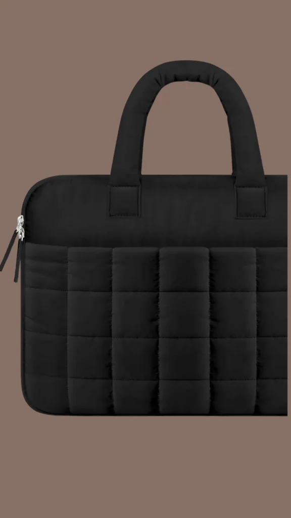 An image from file "2_22.webp" displaying a close-up, partial view of a black padded laptop bag centered against a solid muted brown background. The bag is made of a lightweight, quilted puffer material with a distinct square stitch grid pattern across its lower half. It features two thick, matching black padded top handles stitched securely to the upper body and a silver zipper closure visible on the left side with long fabric pull tabs.
