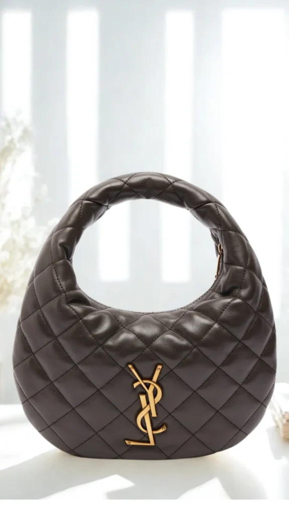 An image from file "2_23.webp" featuring a front-facing view of the Saint Laurent Icarino hobo bag placed on a smooth white surface. The compact, crescent-shaped handbag is crafted from dark brown or soft black quilted leather with a distinct lozenge diamond-stitched pattern that extends smoothly up the integrated curved top handle. A prominent gold metal YSL Cassandre monogram logo is centered on the lower front face of the bag. The background is a brightly lit, soft-focus setting with vertical window pane lines diffusing natural light and subtle white floral accents on the left.
