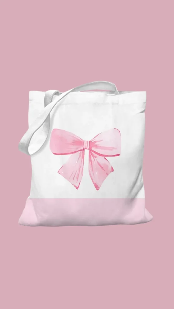 An image from file "2_14.jpg" showing a simple, coquette-style canvas tote bag centered against a solid dusty rose background. The canvas tote is white on the upper portion and features a soft pastel pink color-blocked section along the bottom. Printed prominently in the center of the white canvas is a large, feminine watercolor illustration of a pink ribbon tied into a delicate bow with flowing ends. The tote bag has a simple white fabric shoulder strap draped over the left side.