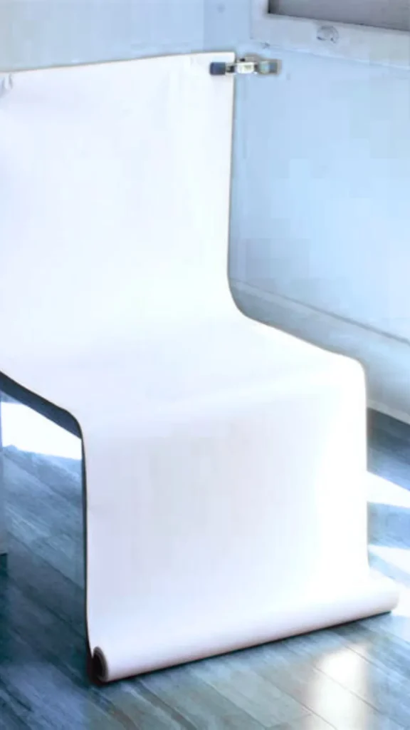 A behind-the-scenes photograph from the file 2_11.webp shows a DIY product photography setup in a brightly lit room with wooden floors. A long roll of thick white paper backdrop is clipped to the top of a chair using a metallic spring clamp. The paper drapes downward across the seat and cascades forward onto the floor in a seamless, curved "sweep" structure to eliminate background harsh angles and corners.