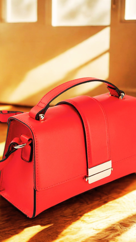 A structured, vibrant red leather crossbody handbag from the file 2_9.png sits on a wooden surface bathed in warm, diagonal sunlight. The bag features a clean, trapezoidal silhouette with a top flat handle attached by gold metal hardware pins. A wide leather vertical band drops down the center of the front flap, securing into a polished, rectangular gold clasp closure at the bottom edge. A matching thin red shoulder strap is attached to the side loop, and the background glows softly with warm, sunlit interior shadows.
