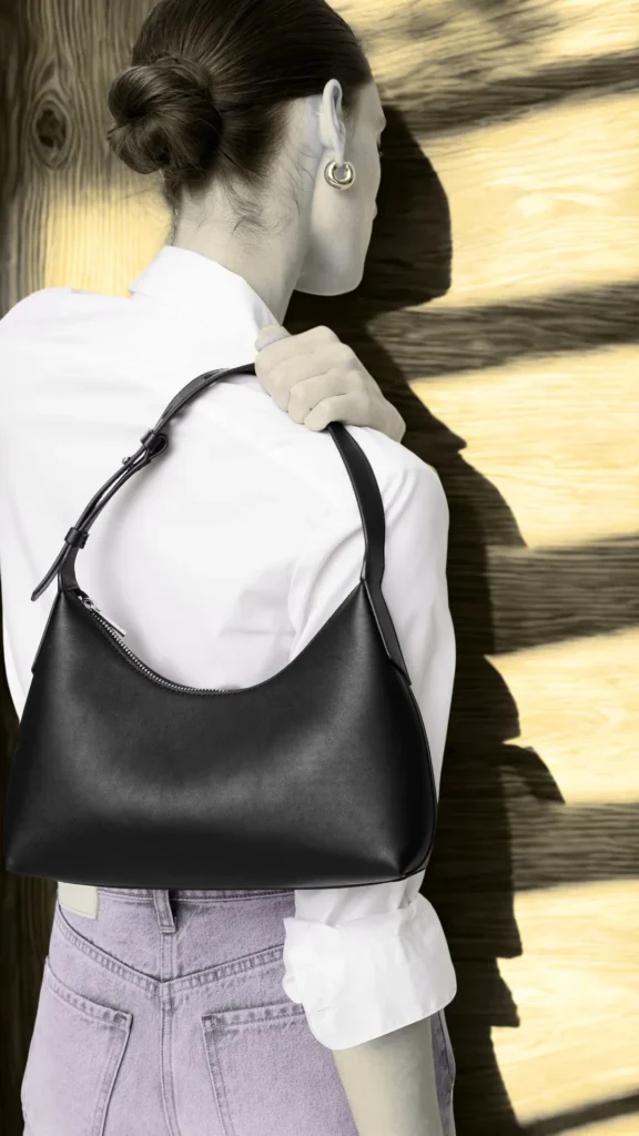 A fashion photograph from the file 2.png shows a woman from behind, with her dark hair styled in a neat bun, modeling a minimalist black crescent-shaped leather shoulder bag. She wears a crisp white button-down shirt, light purple or lavender denim jeans, and small gold hoop earrings. She holds the bag's single adjustable strap over her shoulder. The background features a textured wooden wall highlighted by sharp, angular sunlight and contrasting shadows.