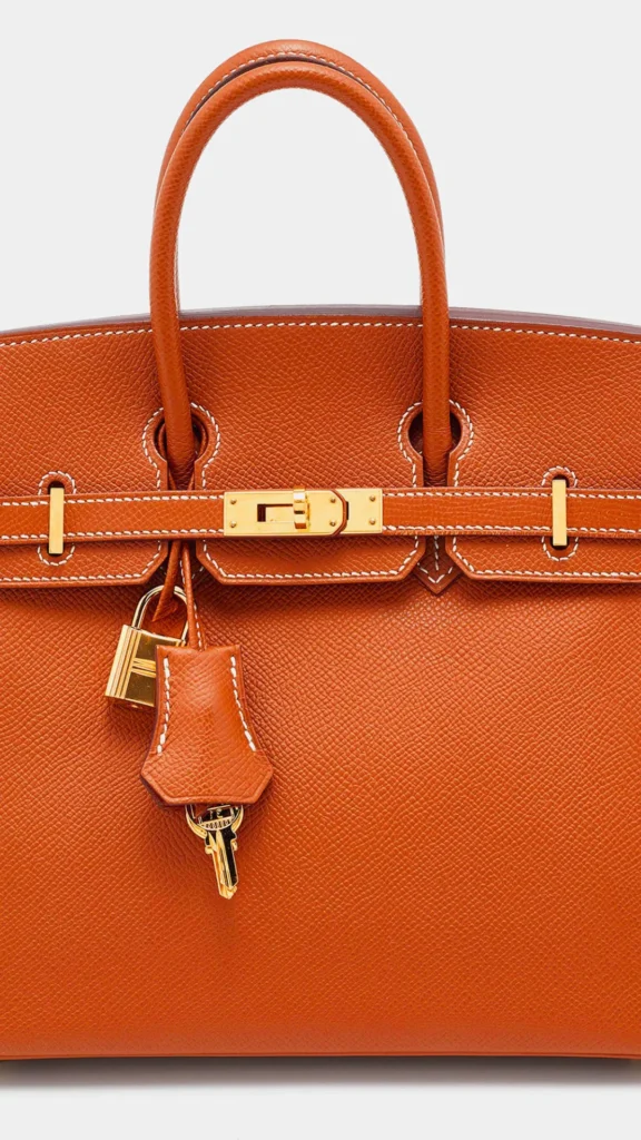 A close-up product photograph from the file 2.jpg highlights the iconic design of an Hermès Birkin or Kelly handbag in textured tan epsom leather. The image details the top handle, contrast white stitching, and gold-plated hardware, including the classic turn-lock closure bar. A matching leather clochette hangs from the handle, holding a small gold padlock and two keys.