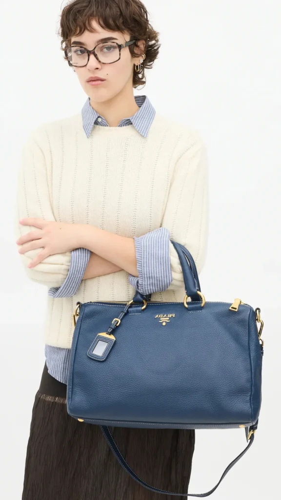 A close-up fashion shot from 3 (1).webp featuring a person with short brown hair and glasses, standing with arms crossed against a white background. They are wearing a cream ribbed knit sweater layered over a blue and white pinstriped button-up shirt. In the crook of their arm, they carry a structured navy blue Prada bowler satchel bag made of durable pebbled leather. The bag showcases prominent gold hardware, a gold logo, and an attached leather luggage tag.