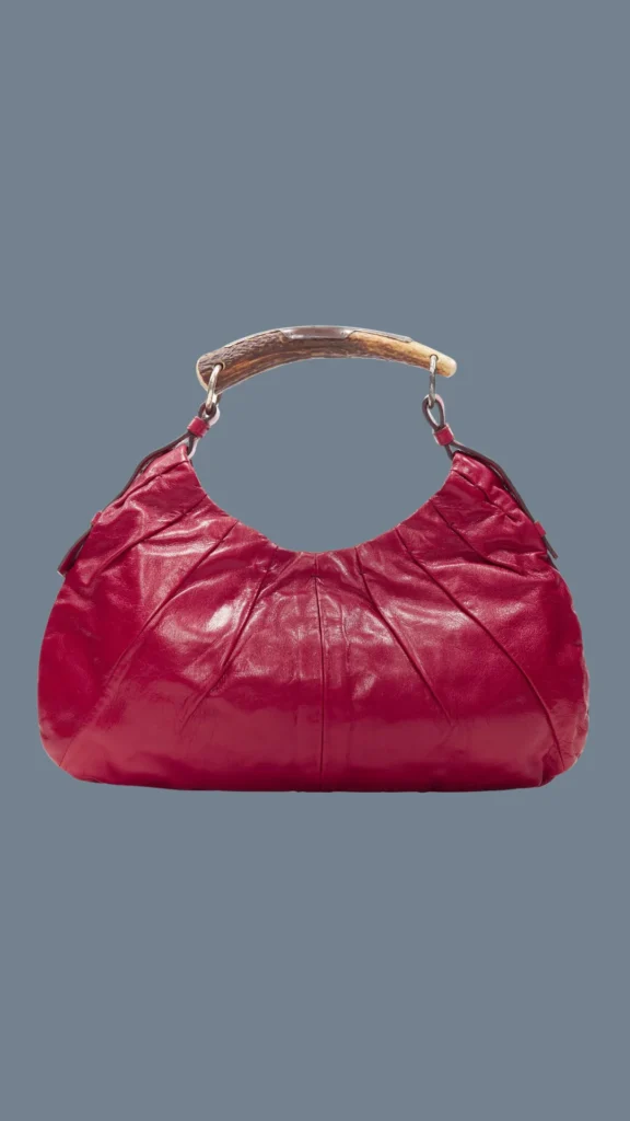 A product shot from 3 (1)_2.webp showcasing a vintage Yves Saint Laurent (YSL) Mombasa hobo bag set against a solid slate blue background. The slouchy pouch handbag is crafted from vibrant berry-red wrinkled leather with elegant vertical pleated paneling. Its most defining feature is the rigid, natural horn-shaped top handle made of polished deer antler, connected to the leather body with silver-tone metal ring hardware.