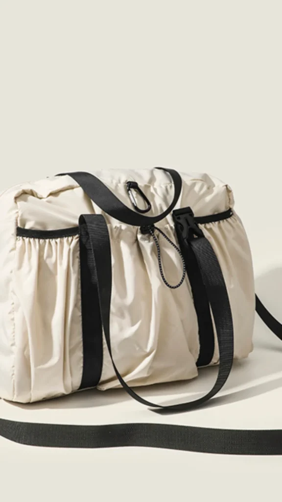 A product shot from 3 (1)_3.webp showcasing a casual, lightweight beige nylon duffel-style bag resting on a cream surface. The slouchy bag features gathered, ruched fabric detailing on the front pocket panels, strongly contrasted by thick black woven straps and handles. It is designed for utility with a black plastic buckle fastener, a drawstring cord, a black carabiner clip, and a long black crossbody shoulder strap laid out in front.