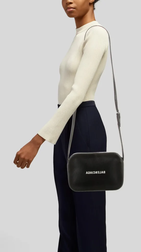 An image from file 3 (1)_3.webp showing a model from the waist up, posed in profile against a neutral grey background. The person is wearing a long-sleeved, ribbed off-white turtleneck top tucked into high-waisted dark navy blue trousers. A rectangular black pebbled-leather camera crossbody bag is slung over their shoulder, resting at the hip. The bag features a silver top zipper and the brand name "BALENCIAGA" printed across the front in white block letters.