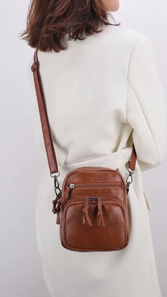 An image from file 3 (1)_4.webp showing a person from a side-back view wearing a structured white blazer and carrying a small, compact brown textured leather crossbody bag. The bag features multiple front zippered compartments with knotted leather tassel zipper pulls and dark metallic hardware connecting the matching brown shoulder strap.