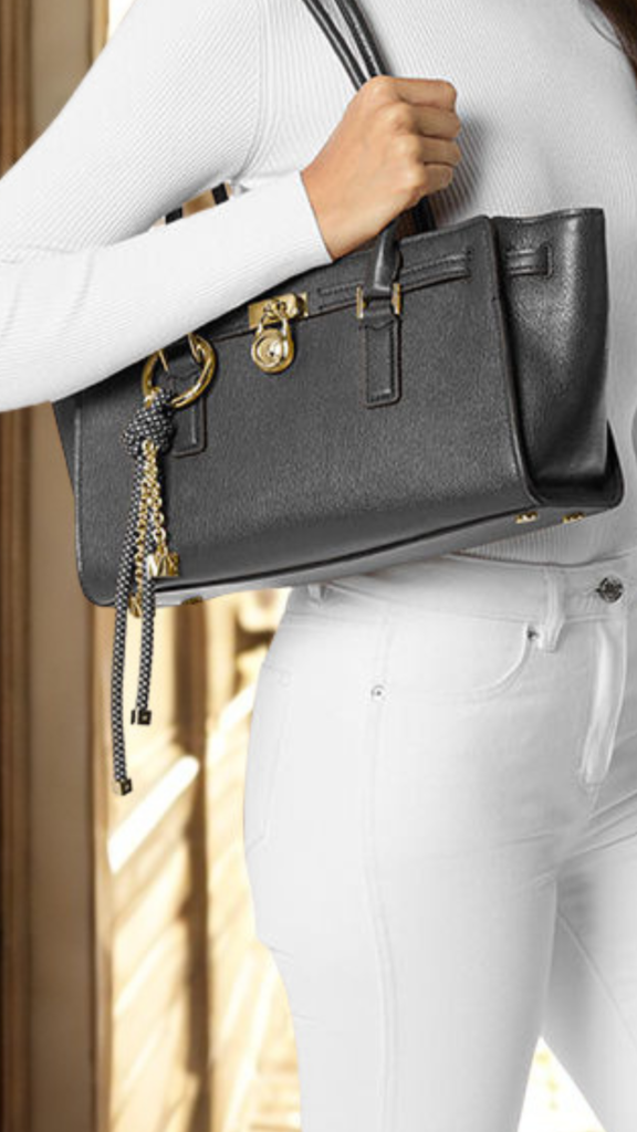 A three-quarter fashion shot from the file 3.png captures a person walking outdoors, modeling a structured black leather satchel bag held by its top handle. The trapezoidal handbag features a clean, smooth texture and a secure front flap design. The person is dressed in a crisp, oversized white button-up shirt paired with a dark, pleated midi skirt. The background reveals a softly blurred street scene with concrete pavement and warm, natural sunlight casting subtle shadows.
