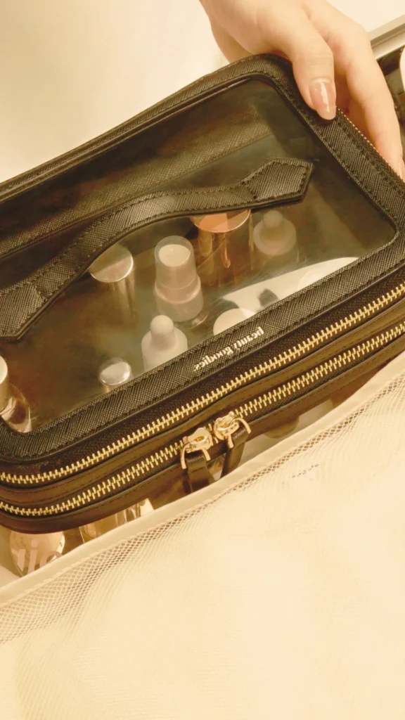 A close-up shot from the file 3_9.webp shows a person's hand with manicured nails lifting a black, rectangular cosmetic travel case. The bag has a clear plastic window on top, revealing several small, upright travel bottles, containers, and skincare products inside. A textured black fabric handle runs across the top window, and a sturdy gold zipper with dual sliders wraps around the perimeter. The scene is bathed in a warm, yellowish lighting, and a white mesh storage netting or fabric is visible along the bottom edge of the frame.