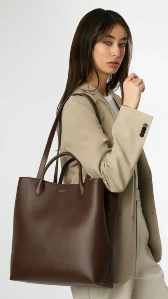 An image from file 3 (1)_6.webp showing a young woman with long, straight brown hair posing against a plain white background. She is wearing a professional, oversized beige blazer over a white top and matching tailored beige trousers. Over her shoulder, she carries a large, structured dark brown leather handbag featuring both dual top handles and a longer shoulder strap. The bag has a minimalist design with clean lines and subtle gold branding near the top rim.