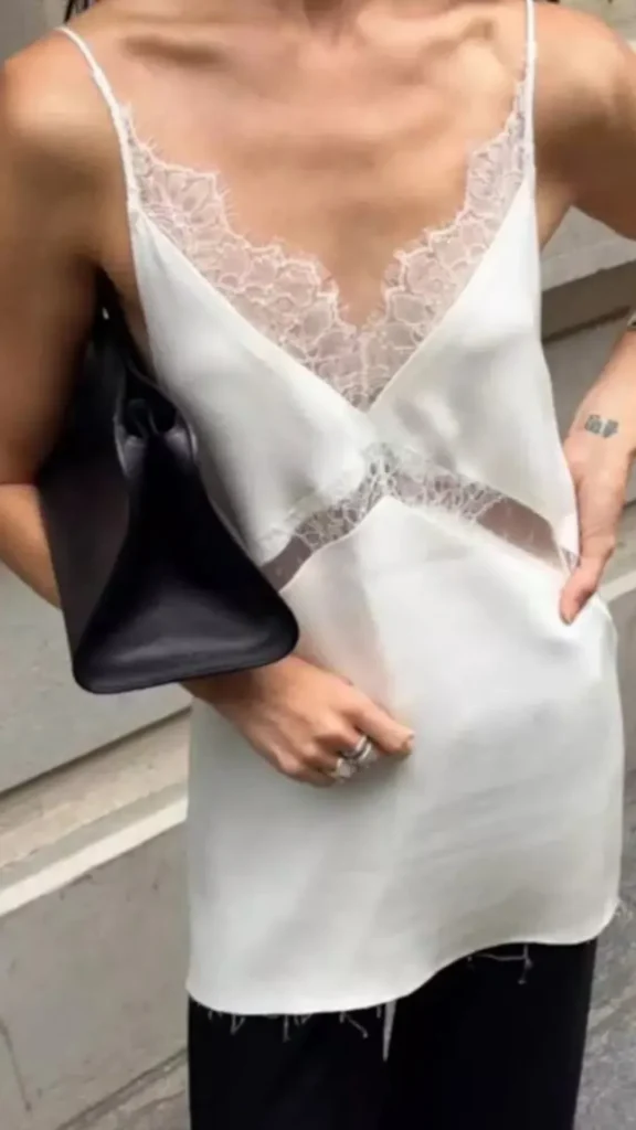 A close-up fashion image from 3.webp showing a person tucked into an effortless outfit. They are wearing a white satin camisole top with intricate lace trim detailing along the V-neckline and midsection, paired with dark pants. Tucked securely under their right arm is a structured, logo-less black leather clutch bag featuring a distinct wide, triangular base profile. The individual wears minimal silver rings on their fingers.