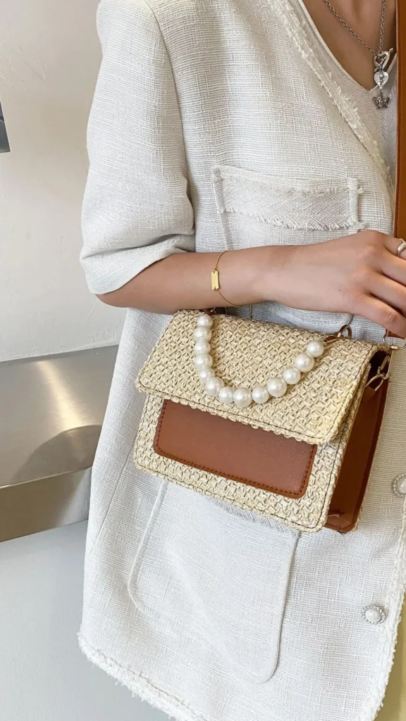 A close-up fashion shot from 3_5.jpg featuring a woman wearing a textured, raw-hem off-white tweed blazer with large pearl buttons, a delicate gold bar bracelet, and a silver necklace. She carries a structured crossbody bag crafted from woven beige straw or raffia material, accented with a smooth tan leather front pocket panel. A short, arched top handle made of large, glossy white pearls rests elegantly across the front flap of the bag.