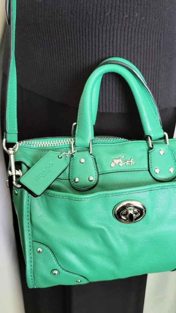 A close-up lifestyle shot from 3_5.webp featuring a vibrant emerald green Coach leather satchel bag being worn over the shoulder against a dark grey knit sweater. The structured bag showcases soft pebbled leather, dual rolled top handles, and a silver horse-and-carriage logo emblem on the front. It is decorated with prominent silver hardware, including stud details, a top zipper, a matching green leather hangtag, and a polished turn-lock closure on the front slip pocket.