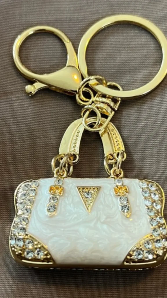 A close-up product photograph from 3_6.webp showing a decorative gold keychain featuring a miniature handbag charm. The tiny handbag charm is made of glossy white enamel with subtle swirls, embellished with micro-rhinestones along the edges and straps. It includes a triangle logo detail and tiny gold top handles, attached to a larger gold lobster claw clasp and split keyring resting against a soft, textured brown fabric background.