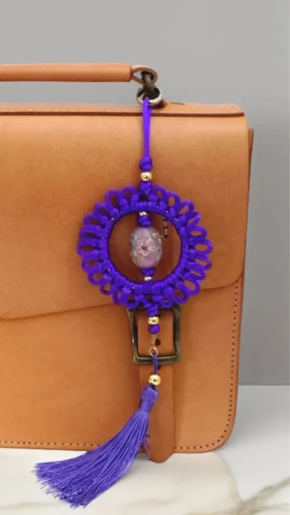 A close-up product photograph from 3_8.webp showing a handmade purple macramé bag charm attached to a structured tan leather satchel. The charm features an intricate, circular woven wheel pattern with looped borders, framing a large, oval-shaped light purple bead with pink floral details suspended in the center. Small gold accent beads decorate the vertical cord above and below the wheel, which culminates in a long, silky purple tassel hanging past the bag's central buckle strap.