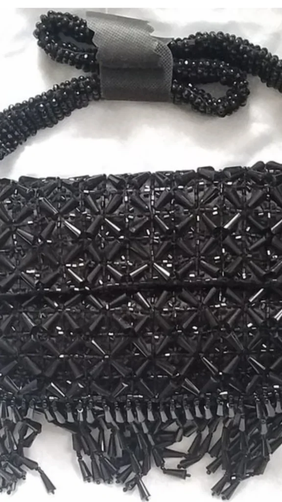 A close-up product photograph from 3_9.webp showcasing an intricate evening clutch bag completely covered in glossy black beadwork. The rectangular flap bag features a geometric pattern made of long, tubular bugle beads and seed beads arranged in starburst clusters. A thick, woven strap made of small black seed beads loops at the top, and rows of dangling black beaded fringe finish the bottom edge, creating texture and a glamorous boho look against a white background.