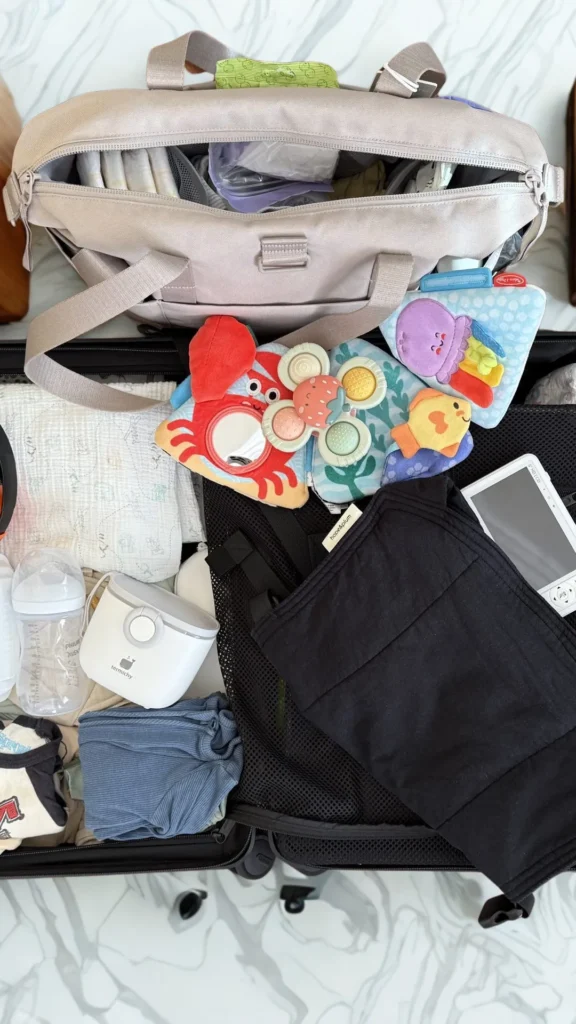 An overhead flat-lay photograph from 3_2.png showing a black suitcase and a beige travel diaper bag being packed with infant essentials. Inside the organized compartments are neatly folded baby clothes, muslin blankets, a clear baby bottle, a white baby food warmer, and a black fabric baby carrier. Scattered on top are bright, colorful sea-themed plush baby toys including an octopus, a fish, a crab with a built-in mirror, and a strawberry-shaped plastic teething toy.
