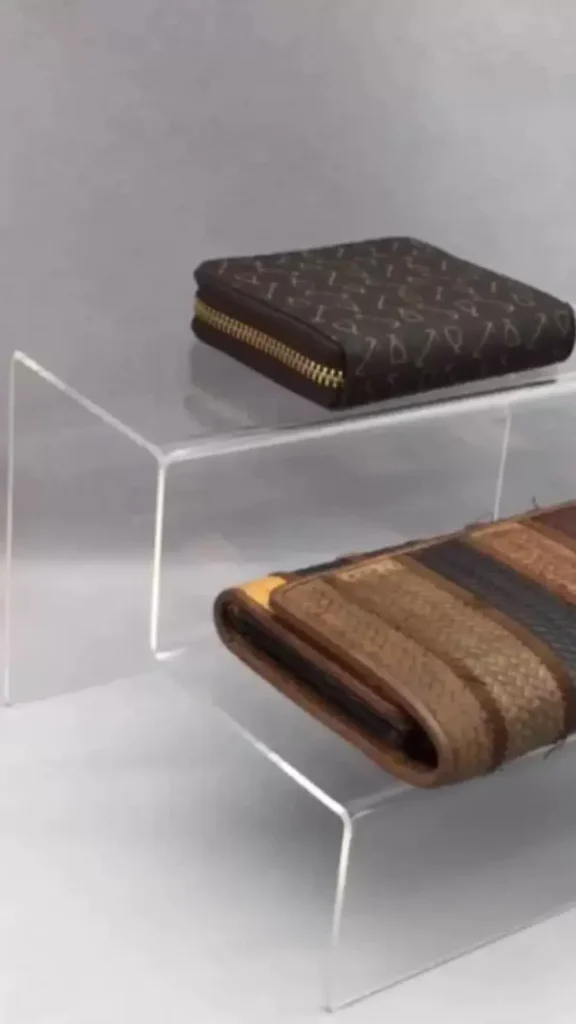 An image from file "3_19.webp" showcasing a clear acrylic tiered riser display stand, perfect for optimizing shelf space. The top tier features a compact, square dark brown patterned wallet with a gold zipper closure. The lower tier displays a larger, brown trifold wallet with multi-textured, woven horizontal panels in different shades of brown and tan. The setup is presented against a neutral grey background, highlighting a boutique-style storage solution.