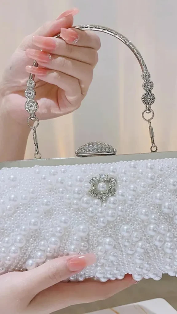An image from file "3_20.webp" showing a close-up, cropped view of an elegant, vintage-style evening clutch bag being held. A person with manicured pink acrylic nails holds the silver metal top handle, which is decorated with textured, circular crystal spacers. The rectangular clutch features a silver metal frame with a rounded rhinestone clasp on top. The exterior of the bag is fully embellished with white faux pearls arranged in an intricate chevron and quilted pattern, with a central floral accent made of sparkling rhinestones and a pearl in the middle.