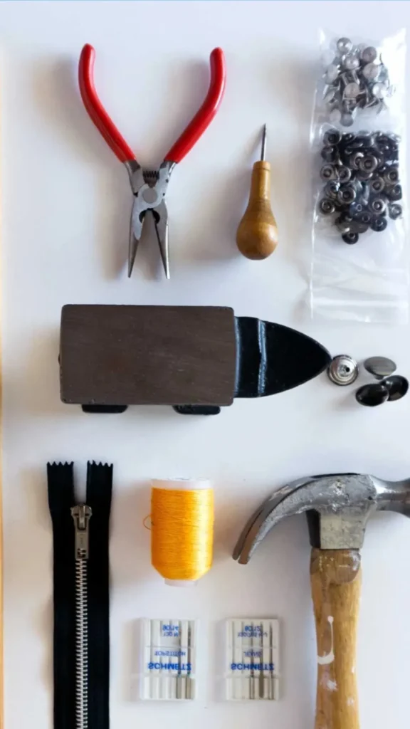 An image from file "3_21.webp" showing a flat-lay arrangement of crafting and sewing supplies used for heavy-duty projects like making a denim handbag, set against a clean white background. The tools include a pair of pliers with red handles, a wooden-handled awl, two small plastic bags filled with silver metal snaps or rivets, a heavy rectangular metal block or anvil piece, a black metal zipper, a spool of thick bright yellow thread, a traditional claw hammer with a wooden handle, and two small rectangular packs of sewing machine needles.