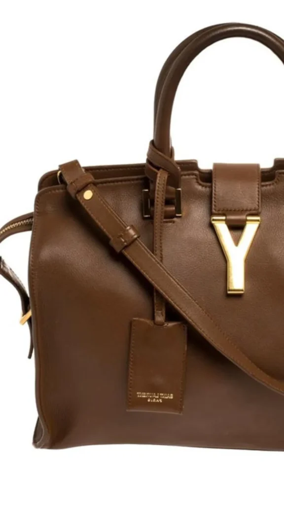 An image from file "3_23.webp" displaying a cropped, close-up view of a classic brown leather luxury tote bag, identified as a Saint Laurent design. The structured handbag is crafted from smooth, rich brown leather and features dual rolled top handles, an attached adjustable leather shoulder strap, and a hanging leather clochette or ID tag with subtle gold embossed branding. A prominent, polished gold metal "Y" hardware emblem serves as a striking clasp feature, securing the small leather tab closure at the top center of the bag's front panel. The image is set against a plain, clean white background.