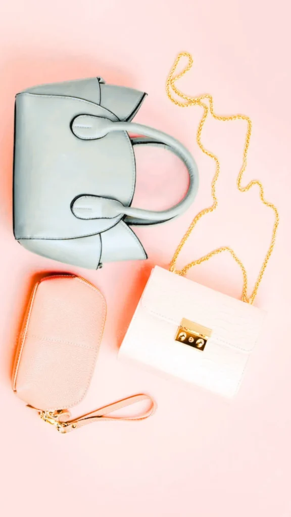 A top-down flat lay shot from the file 3_10.webp displays three small fashion accessories arranged on a solid pink background. At the top sits a light blue-grey structured handbag with rounded double top handles and winged side panels. Below it on the right is a small, structured white crossbody purse with a faux-croc embossed texture, a rectangular gold clasp closure, and a long gold chain strap. To the left is a small pink pebbled-leather cosmetic pouch or wristlet with a gold zipper and a wrist strap attached.