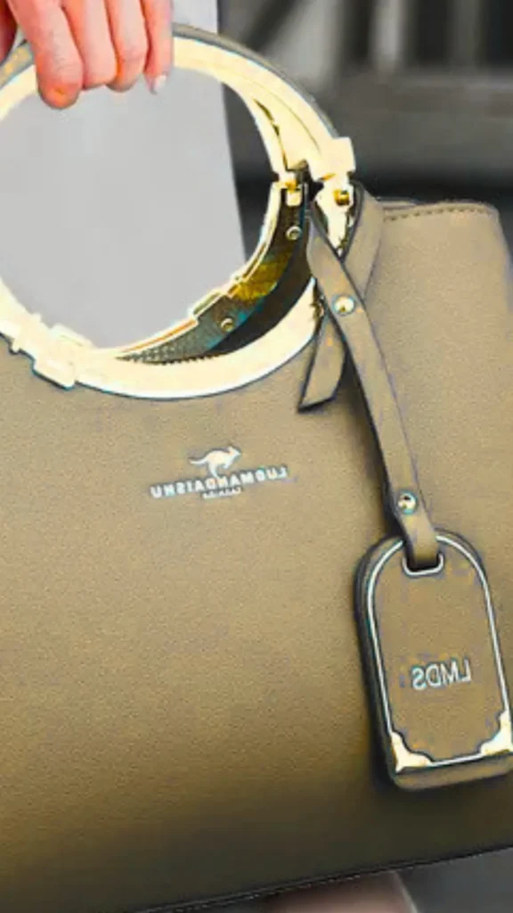 A close-up fashion photograph from the file 1.png shows a person holding a structured, trapezoidal handbag in a rich, warm brown color. The bag features a classic design with structured top handles, gold feet visible at the base, and a matching leather strap with a gold buckle detailing wrapped around the upper frame. The person carrying the bag wears a gold statement ring and light-wash blue denim jeans.