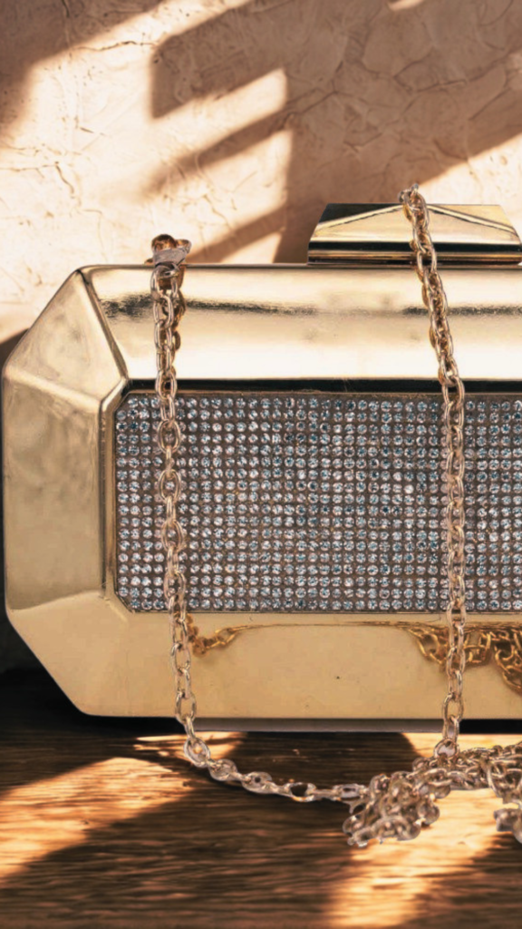 A close-up shot of a glamorous, metallic gold box clutch handbag from the file 3_10.png resting on a wooden surface. The evening bag features a structured, octagonal silhouette with thick, beveled gold borders framing a rectangular center panel packed with rows of sparkling, inlaid crystals. A matching gold link chain shoulder strap drapes loosely across the front of the bag, and a geometric gold clasp sits on top. Warm, textured sunlight and leaf shadows are cast on the light-colored wall behind it.