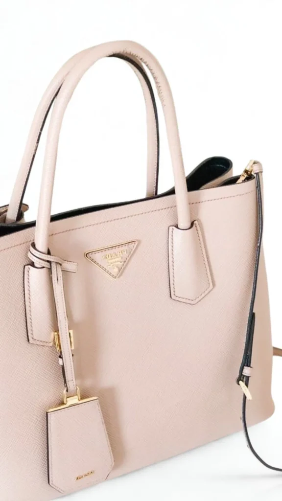 A close-up product photograph from the file 3.webp highlights a luxury Prada Galleria tote handbag crafted from blush-pink or beige cross-grained Saffiano leather. The bag features two structured top handles, side snap closures, and a matching leather key ring tag hanging from one of the handle bases. The signature gold-toned inverted triangle Prada logo plaque is prominently displayed on the front center panel.
