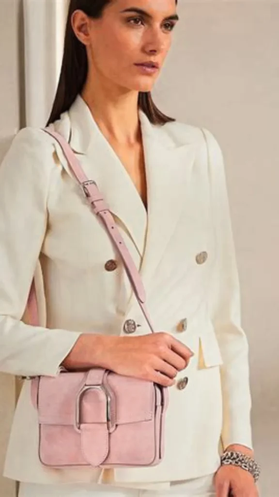 A close-up product photograph from the file 3.webp features a luxurious, structured top-handle handbag in a sophisticated dusty rose pink leather. The bag displays high-end craftsmanship with clean, white stitching along its edges and seams. It has two curved leather handles securely attached to the front panel with stitched tabs, offering a polished look for a professional capsule collection.