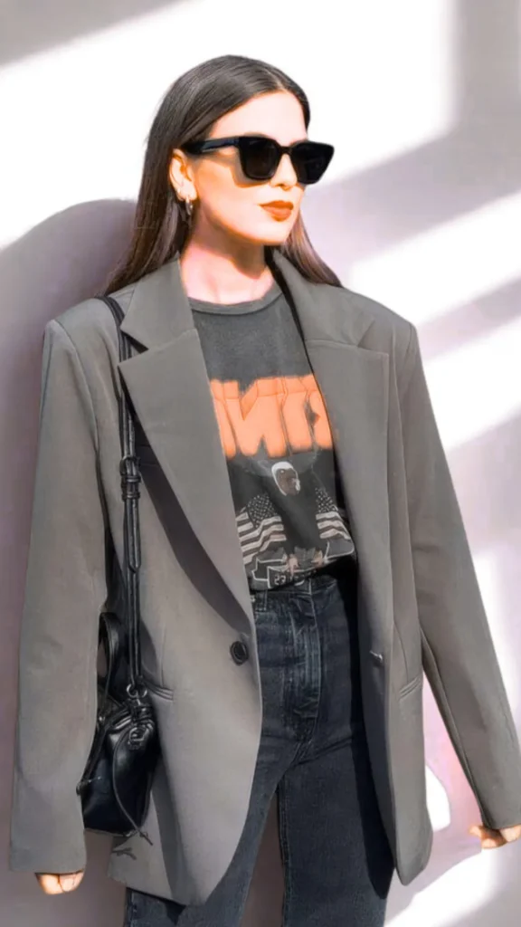 A three-quarter length fashion photograph from the file 3.webp shows a woman walking outdoors in bright sunlight. She wears an oversized, unstructured olive-gray blazer over a dark graphic t-shirt tucked into high-waisted black jeans. She is accessorized with sharp, black cat-eye sunglasses and has a small, black leather crossbody bag slung over her shoulder, hanging subtly at her side against the jacket.