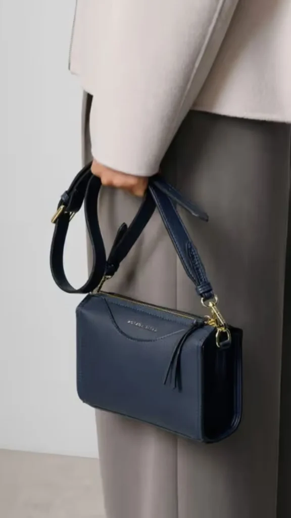 A close-up fashion shot from 4 (1)_2.webp of a person holding a small, structured dark navy blue leather camera-style crossbody bag. The rectangular handbag features a top zip closure with gold hardware, a curved stitch detailing on the front panel, and a thick matching navy leather shoulder strap bunched in their hand. The individual is wearing a light off-white or cream jacket paired with smooth grey trousers, showcasing a distinctive and elegant neutral color palette.