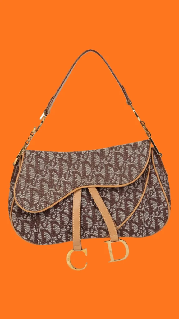 A product shot from 4 (1)_3.webp showcasing a vintage Christian Dior Saddle bag set against a vibrant solid orange background. The bag features the signature brown and beige Oblique monogram canvas pattern with tan leather piping and trim. It is styled with its iconic asymmetrical saddle-shaped front flap, dangling gold-tone metal "C" and "D" logo charms, and a matching shoulder strap connected by gold "CD" hardware links.