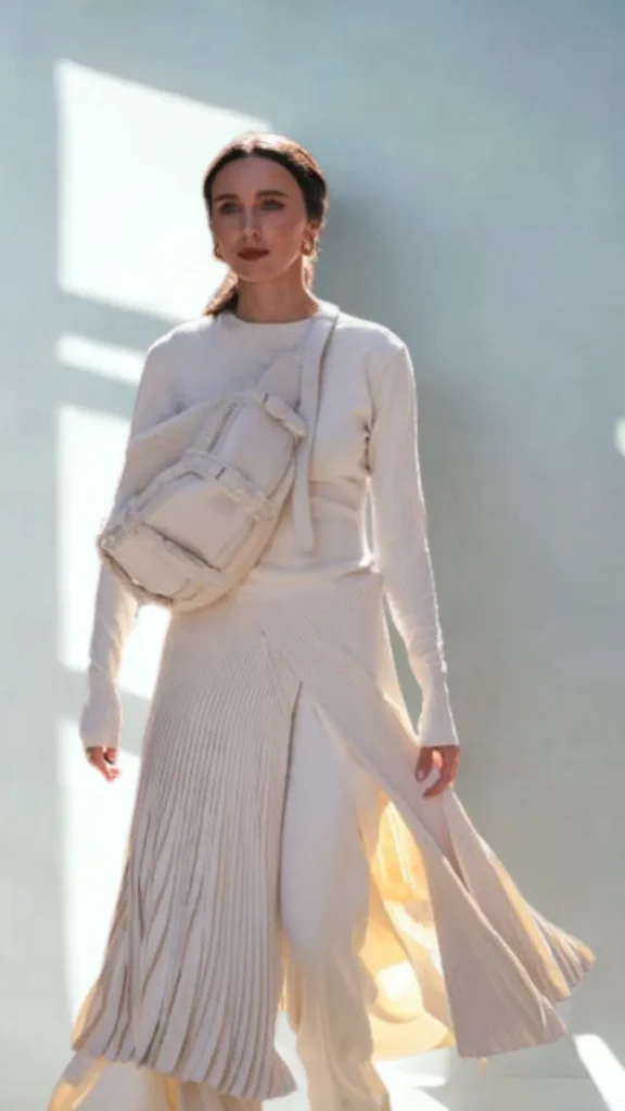 A full-length fashion shot of a woman walking forward in 4 (1)_4.webp, showcasing an elegant monochrome cream outfit. She is wearing a fitted, long-sleeved cream top and a dramatic, flowing asymmetric pleated maxi skirt layered over matching trousers. Worn diagonally across her torso is a large, structured cream-beige utility sling bag with multiple padded compartments and tactical strap details. The background features crisp sunlight and sharp geometric shadows against a light neutral wall.