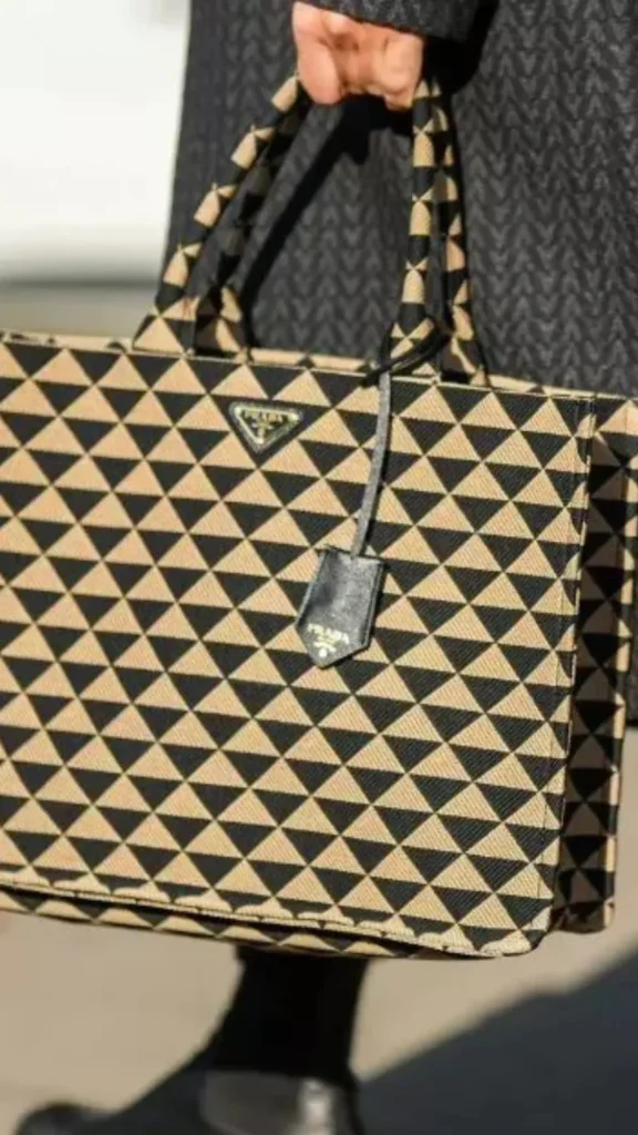 An image from file 4 (1)_3.webp showing a close-up, street-style shot of someone holding a large Prada Symbole tote bag by its top handles. The structured, rectangular fabric bag features a striking, repeating geometric pattern of beige and black triangles. Attached near the top handle is a small black leather clochette or luggage tag, and the iconic triangular Prada logo plaque is visible near the upper rim.