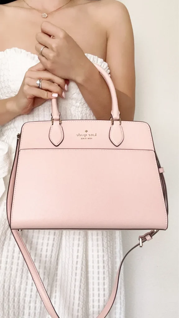 An image from file 4 (1)_4.webp showing a woman in a white textured, strapless dress holding a structured light pink Kate Spade satchel bag. She holds the two rigid top handles with both hands, showcasing her rings and light manicure. The elegant, pastel pink bag features a gold spade logo on the front, a flat slip pocket, and a matching thin pink shoulder strap hanging down.