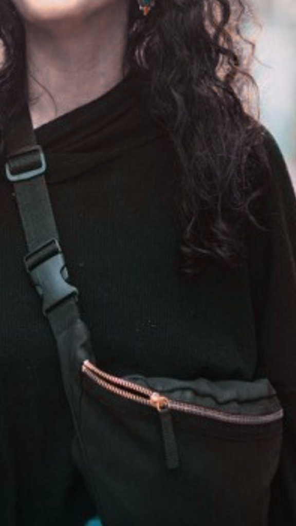 A close-up fashion photograph from the file 4.jpg shows a person's upper body wearing a black cowl-neck top and modeling a utilitarian black canvas sling bag worn across the chest. The compact belt bag features a textured black strap with a heavy-duty plastic buckle closure on the shoulder and a prominent rose-gold metal zipper on the main compartment. The background shows a dimly lit outdoor setting with warm, golden ambient lighting reflecting off the person's long, curly dark hair.