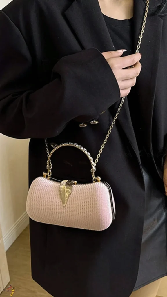 An image from file "4 (1)_6.webp" showcasing a close-up of a small, elegant hard-case evening clutch. The purse features a light pink, textured ribbed exterior with a curved silhouette. It is decorated with an ornate gold leaf-shaped clasp closure at the top, a matching semi-circular gold top handle, and a thin gold chain shoulder strap draped over the user's shoulder. The person holding the bag is wearing a sophisticated black blazer jacket over a black top and a black leather-like skirt.