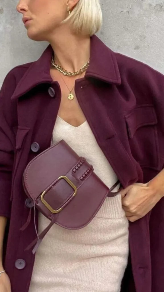 A close-up fashion shot from 4.jpg of a person wearing a cream-colored knit top and matching ribbed skirt. They are layering with an open, oversized dark maroon wool shacket jacket featuring large buttons and chest pockets. Slung across their front is a smooth maroon leather crossbody bag with a large gold buckle, completed with layered gold chain necklaces.