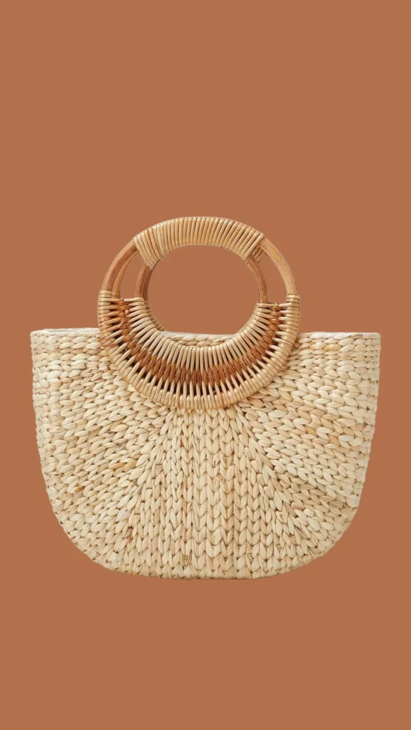 A studio product photograph from 4_4.webp showing a half-moon shaped tote bag woven from light beige natural straw or raffia. The bag features an intricate chevron-style weave pattern across its body. It is designed with prominent, integrated round top handles made of smooth, light-colored bamboo or rattan wrapped in matching natural fibers at the base. The entire handbag is centered against a solid, warm terracotta brown background.