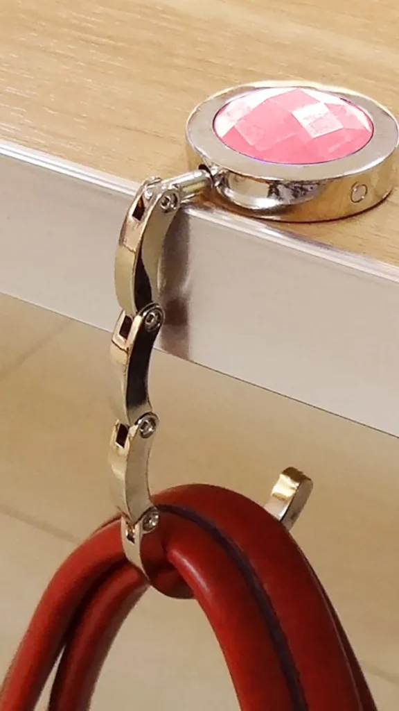 A close-up product photograph from 4_6.webp demonstrating a folding handbag hook in use. The top part of the circular, silver-toned metal hook features a faceted pink gem inlay and rests flat on a light wood-grained table edge with a silver metallic rim. An articulated, segmented metal arm extends downwards from the base, forming an adjustable hook that securely suspends the brown leather handles of a purse, keeping it safely off the floor.