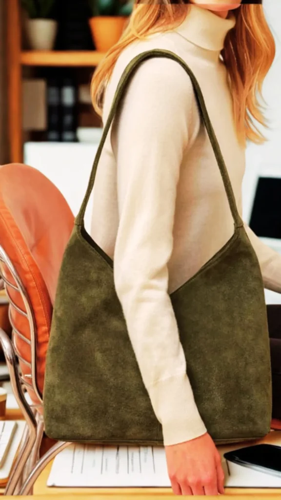 A medium, cropped shot from the file 4_11.png shows a person with blonde hair wearing a cream-colored turtleneck sweater. The individual is modeling a large, slouchy olive-green suede hobo bag carried over the shoulder by its single integrated strap. The setting appears to be an office or creative studio workspace, with the person standing next to a modern tan leather desk chair. Papers, a smartphone, and blurred bookshelves with potted plants are visible in the background.