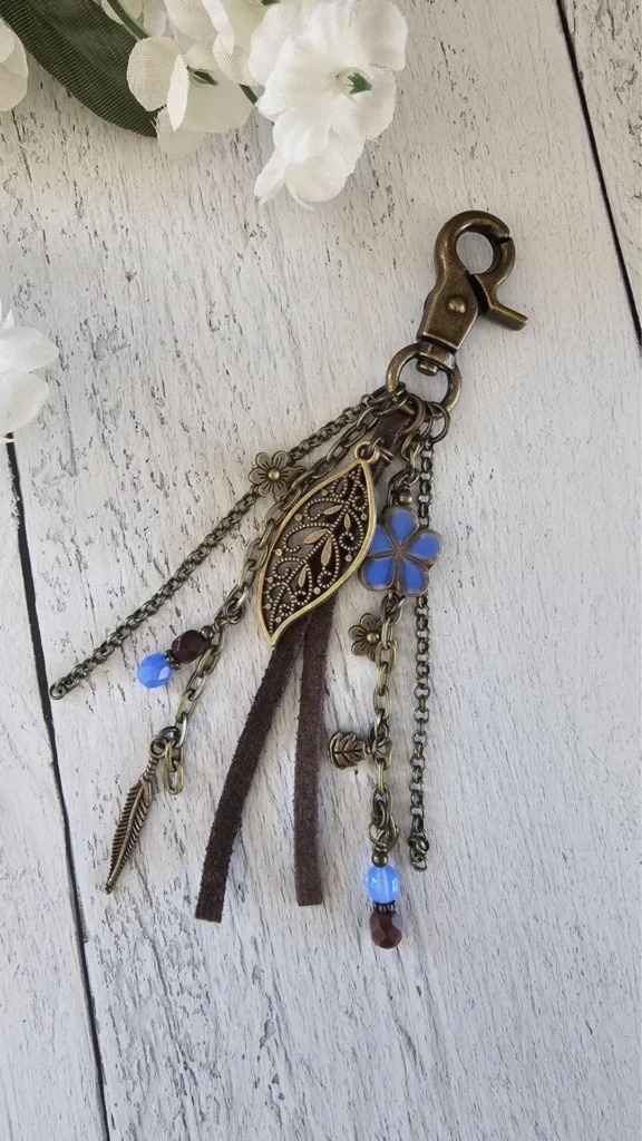 A flat-lay product photograph from 4_4.jpg showcasing a bohemian-style bag charm resting on a white-washed wooden plank surface. The accessory features an antique bronze-toned lobster claw clasp attached to multiple dangling elements, including thin bronze chains, dark brown suede leather strips, and intricate metal pendants like a filigree leaf, a feather, and tiny flowers. Accent beads in light blue and dark brown add a subtle pop of color, with white artificial flowers softly framing the top corner.