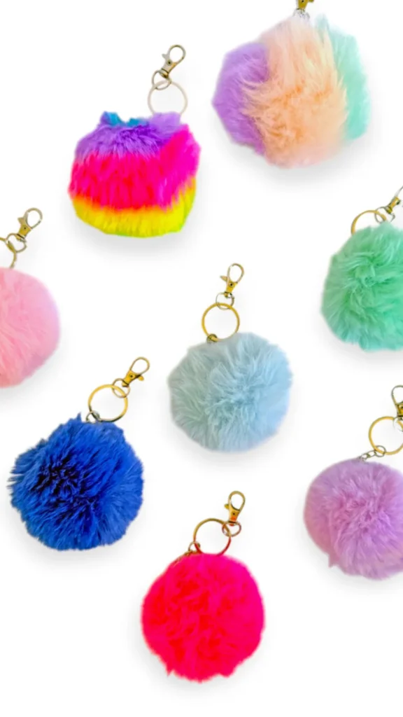 A flat-lay product photograph from 4_7.webp displaying an assortment of fluffy faux-fur pom-pom bag charms scattered across a clean white background. Each soft keychain features a dense, round pom-pom attached to a gold split ring and a matching small lobster claw clasp. The collection includes a mix of solid colors—like hot pink, deep royal blue, lavender, pastel mint green, bubblegum pink, and ice blue—as well as two multi-colored variants showing rainbow and soft pastel gradients.